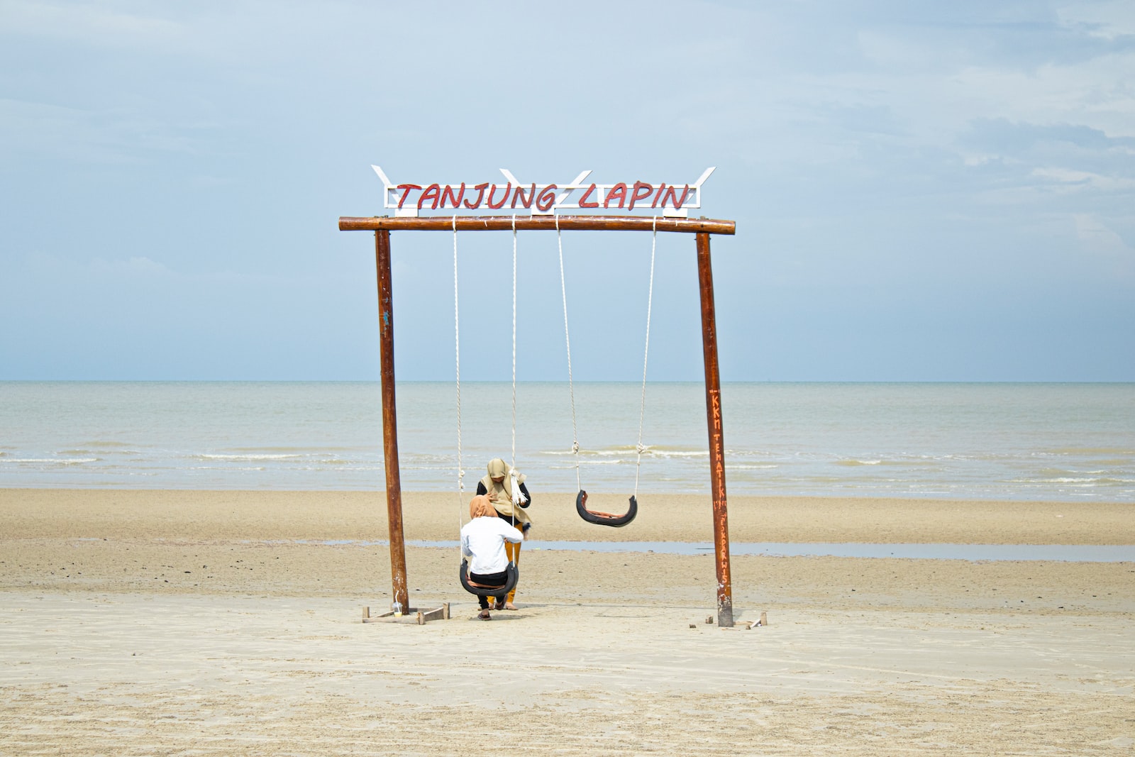 man in white t-shirt and black pants sitting on brown wooden swing chair on beach